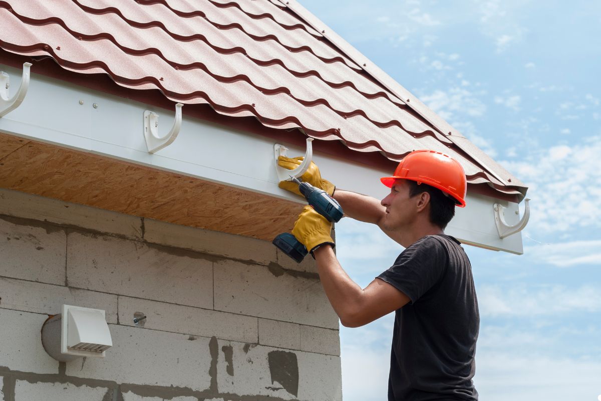 A man operates a drill while installing a gutter on a house, focused on his task with tools nearby.